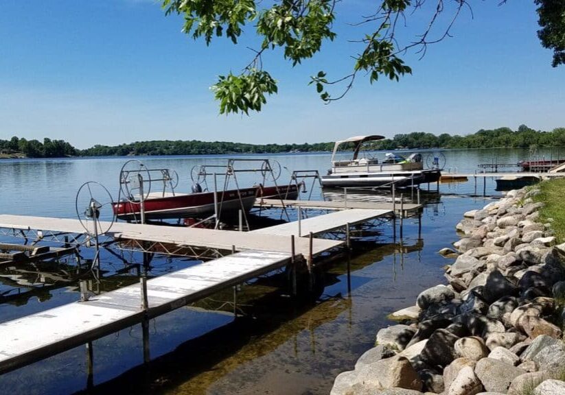 A peaceful lakeside scene at Vacationer's Inn, with docks extending over the water, two boats moored, and a rocky shoreline bordered by green grass under a clear blue sky. Trees are visible in the distance across the lake.