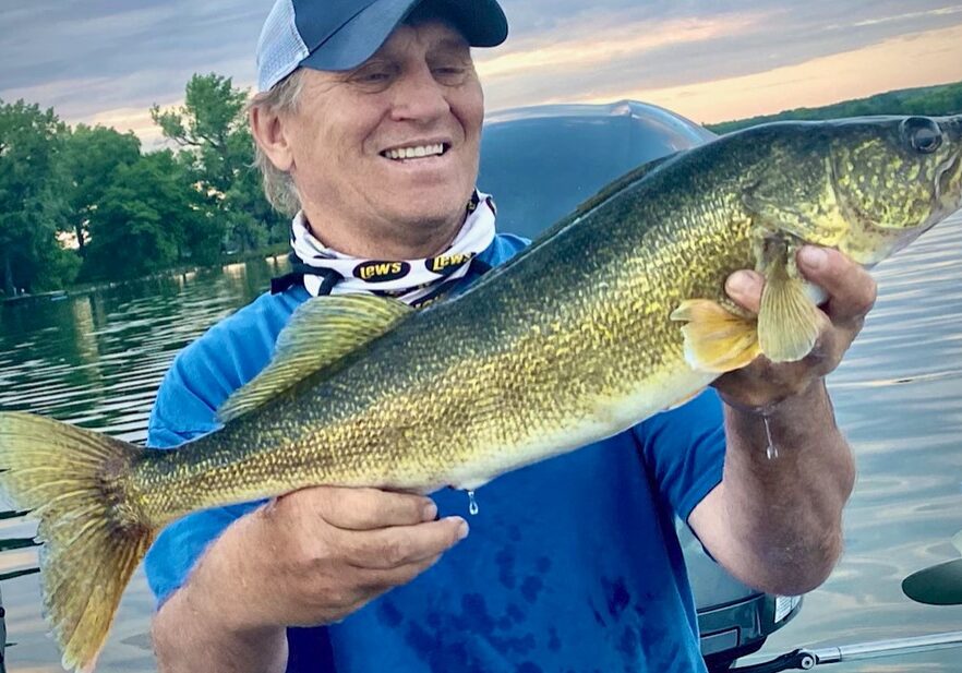 A smiling man in a blue shirt and cap proudly holds a large, freshly caught walleye in both hands while standing on a boat on a calm lake, with trees and a cloudy sky in the background.