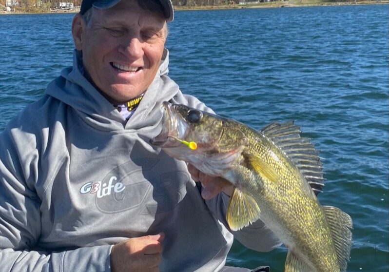 A smiling person in a gray hoodie and cap holds a large yellow-green fish with a lure in its mouth while enjoying perfect fall fishing weather from a boat on a blue lake. Trees with autumn leaves are visible in the background.
