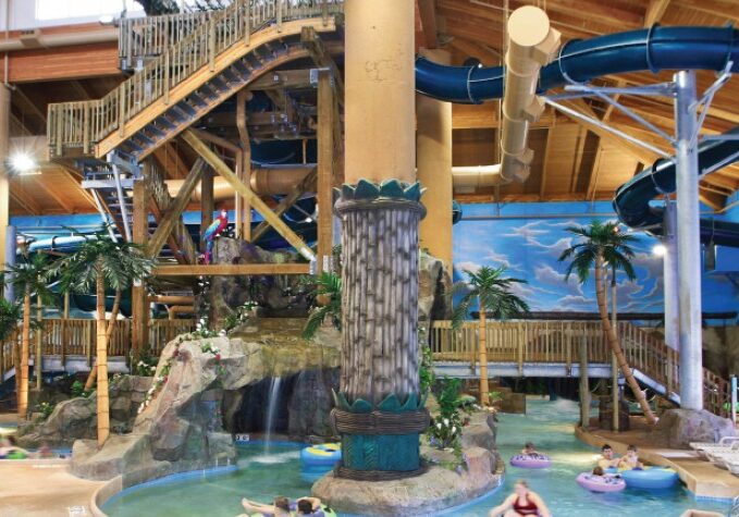 Big Splash Indoor Waterpark features a large blue waterslide, wooden walkways, fake palm trees, and people floating in inner tubes. Bright, tropical-themed decor and a high wooden ceiling complete the fun atmosphere.