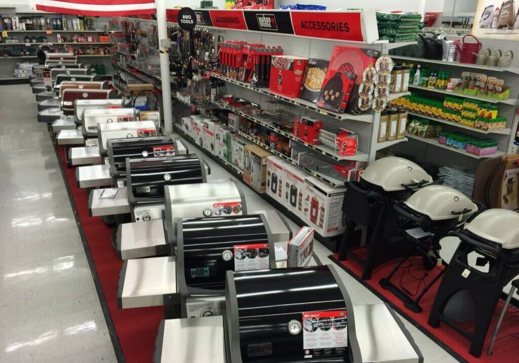A store aisle at Ace Hardware displays various grills in the foreground, with shelves of grilling accessories, tools, and lawn and garden products in the background. The section is well-lit and organized—perfect for your next visit to an Ace Hardware store near me.