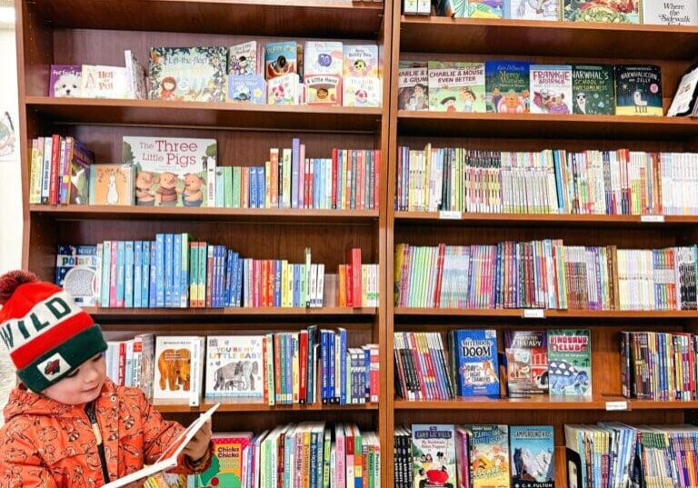 A young child in an orange jacket and winter hat stands in front of tall bookshelves filled with colorful children’s books, enjoying an open book during Saturday Story Hour.