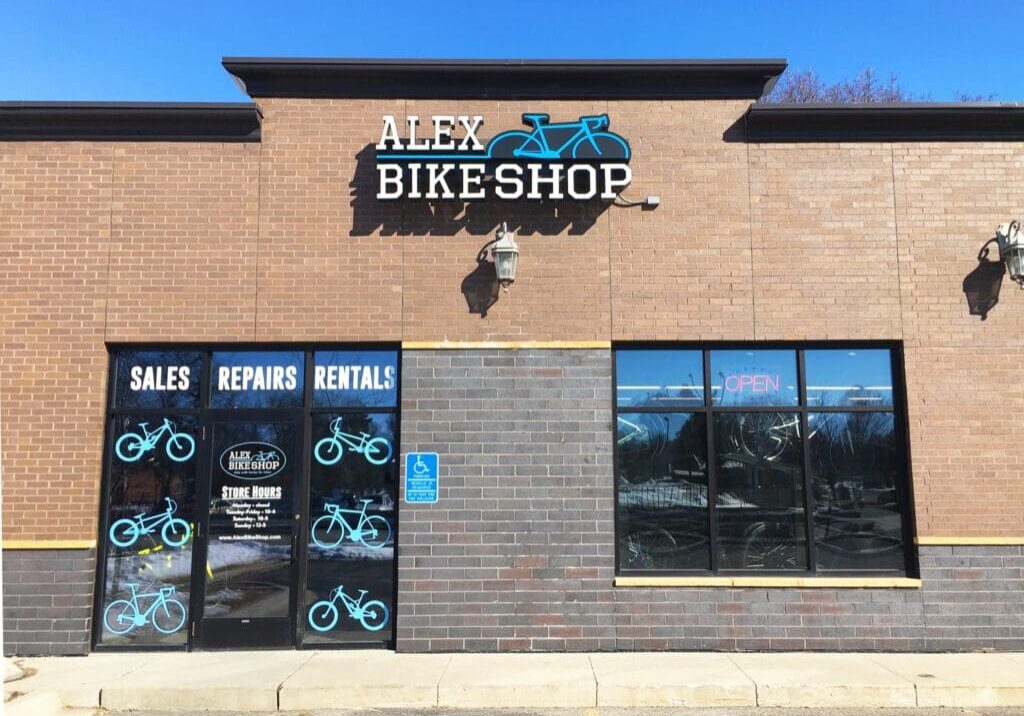 A brick storefront for Alex Bike Shop features a blue bicycle logo. Windows display bike graphics and the words “Sales, Repairs, Rentals.” An “Open” sign is lit inside, and there’s a handicap parking sign by the door.