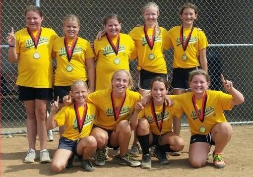 Nine girls in yellow "Athletics" shirts and medals pose on a dirt field, smiling and holding up one finger, proudly representing Alexandria Sports & Activity Organizations after winning a game or tournament.
