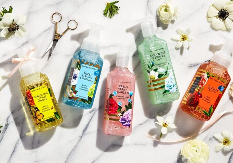 Five colorful hand soaps with floral labels, including Bath & Body Works, are arranged on a white marble surface, surrounded by white flowers, gold scissors, and a pink ribbon. Scents featured include peach and cucumber.