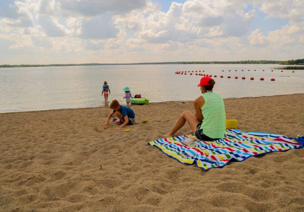 A woman sits on a colorful striped blanket on a sandy beach at Lake Le Homme Dieu near Alexandria, MN, watching two children play in the sand while another stands at the water’s edge under a partly cloudy sky.