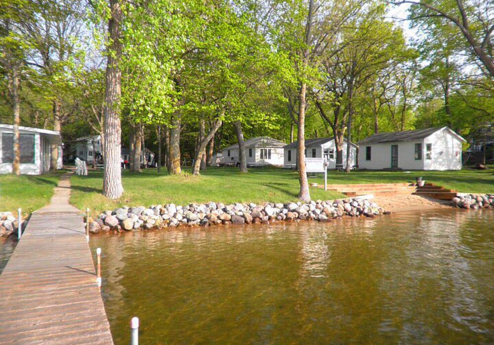 A wooden dock extends into a clear lake at Berg's Resort, with a sandy shore. Several white cottages are nestled among tall, leafy trees, and green grass and rocks line the shoreline on a sunny day.