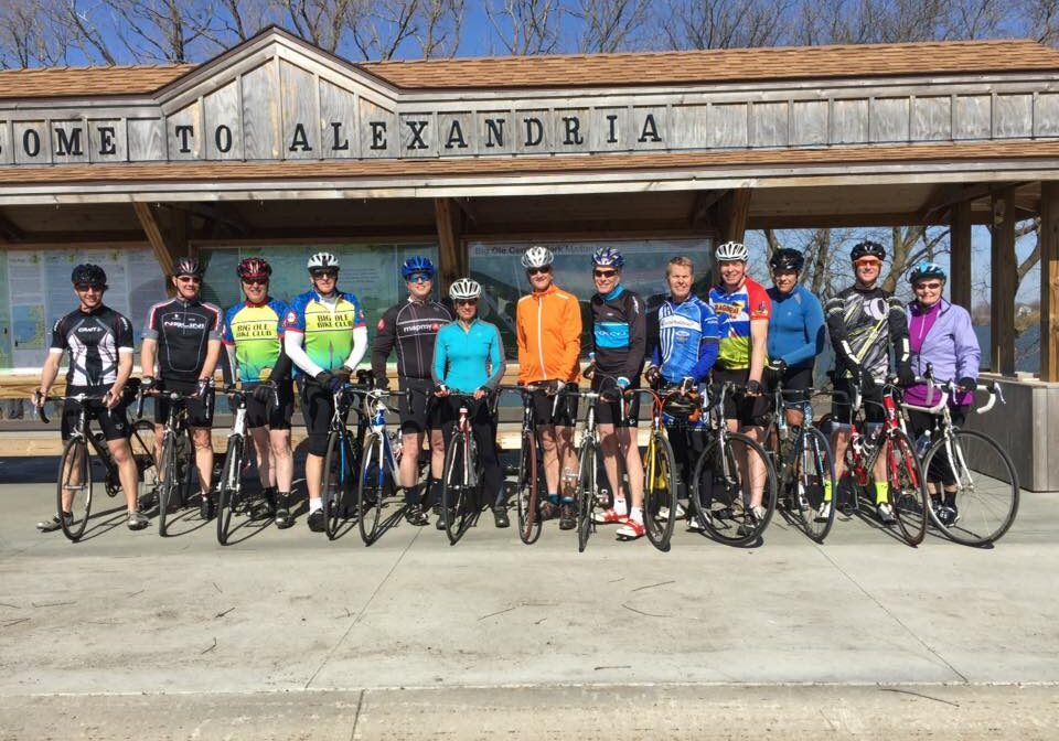 A group of 13 cyclists from the Big Ole Bike Club, wearing helmets and cycling gear, stand with their bicycles in front of a wooden shelter that says "Welcome to Alexandria" on a sunny day.