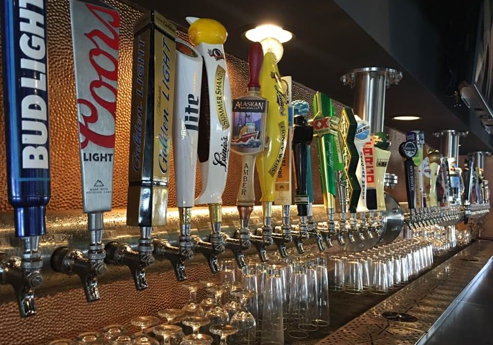 A row of beer taps with colorful handles, each labeled with different beer brands, lines the Boulder Tap House bar above upside-down hanging glasses and a counter ready for serving drinks.