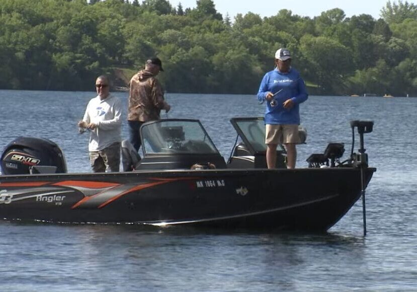 Three people are fishing on a black motorboat with orange stripes on a calm lake, surrounded by lush trees and greenery along the shore.