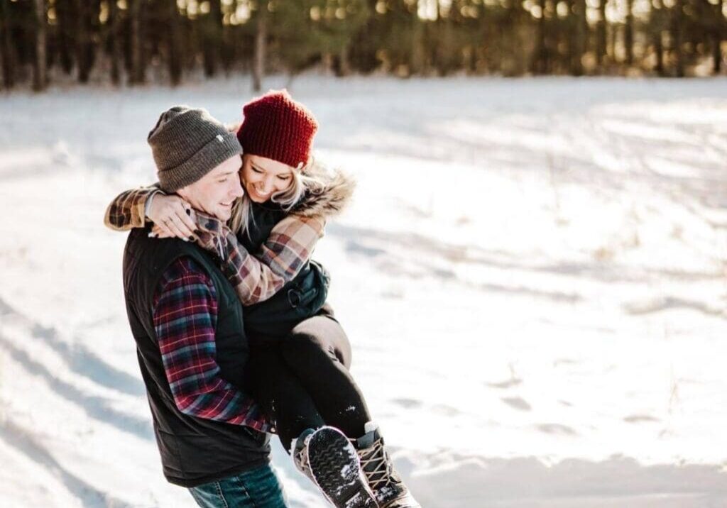 A man in winter clothes lifts a smiling woman wearing a red knit hat and boots in a snowy Alexandria MN field with pine trees behind them. Sunlight shines through, creating a warm, cheerful Valentine’s Day atmosphere.