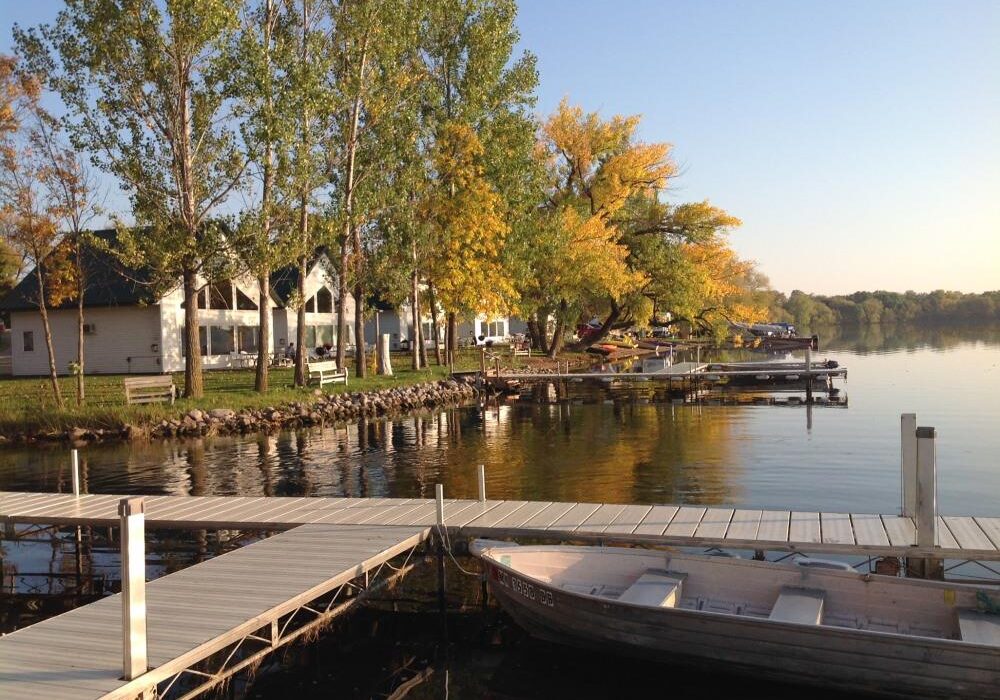 A calm lake with wooden docks and a rowboat in the foreground at Brophy Lake Resort, lined with trees showing green and yellow leaves. Houses and benches sit along the waterfront under a clear sky.