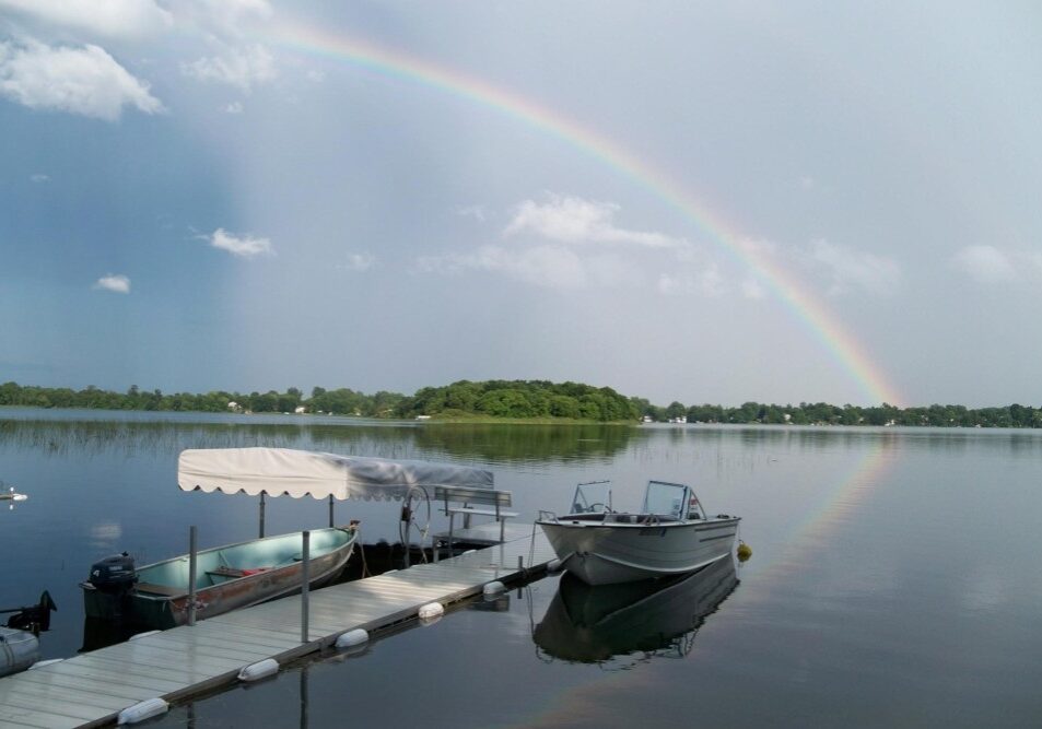 A rainbow arcs across a calm lake with a dock, several small boats, and an island of trees in the background. Enjoy this peaceful scene—and consider boat rentals at Brophy Lake Resort for your own relaxing getaway.