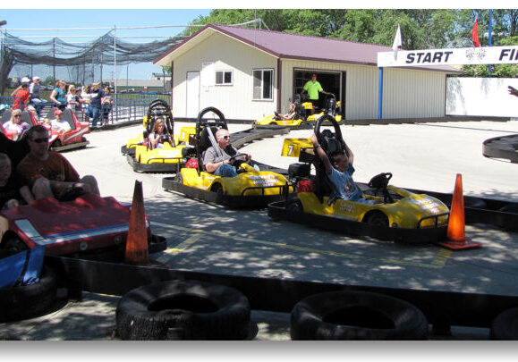 People are riding go-karts at Casey's Amusement Park on an outdoor track near the start/finish line, with a few spectators watching in the background. Some drivers are waiting in yellow karts while a worker in a green shirt stands nearby.