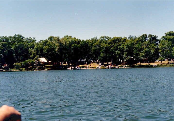 A calm lake with gentle ripples in the foreground, bordered by a tree-lined shore in the background at Chippewa Park. People can be seen near the water's edge, enjoying a sunny day.