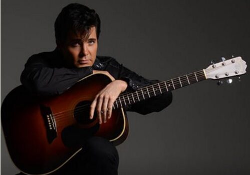 A person with dark hair, dressed in black, sits holding an acoustic guitar across their lap against a plain dark background. The individual, a performer for the Central Lakes Concert Association, looks directly at the camera with a serious expression.