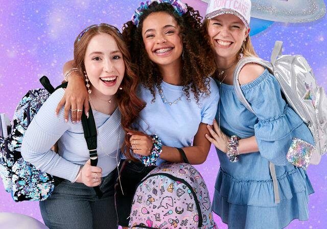 Three smiling girls with backpacks stand together in front of a purple, outer-space-themed background with planets. Wearing pastel and sparkly Claire's clothes and accessories, they appear happy and excited.