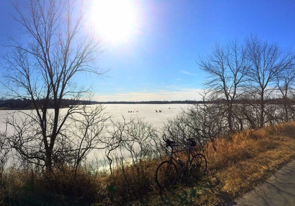 A bicycle rests on a sunlit path in Alexandria beside leafless trees, overlooking a frozen lake where people ice fish beneath a clear blue sky as spring arrives with clear trails.