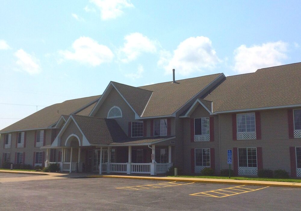 A two-story Quality Inn with a large porch, beige siding, red-framed windows, and a gabled roof sits in front of a mostly empty parking lot under a partly cloudy sky.