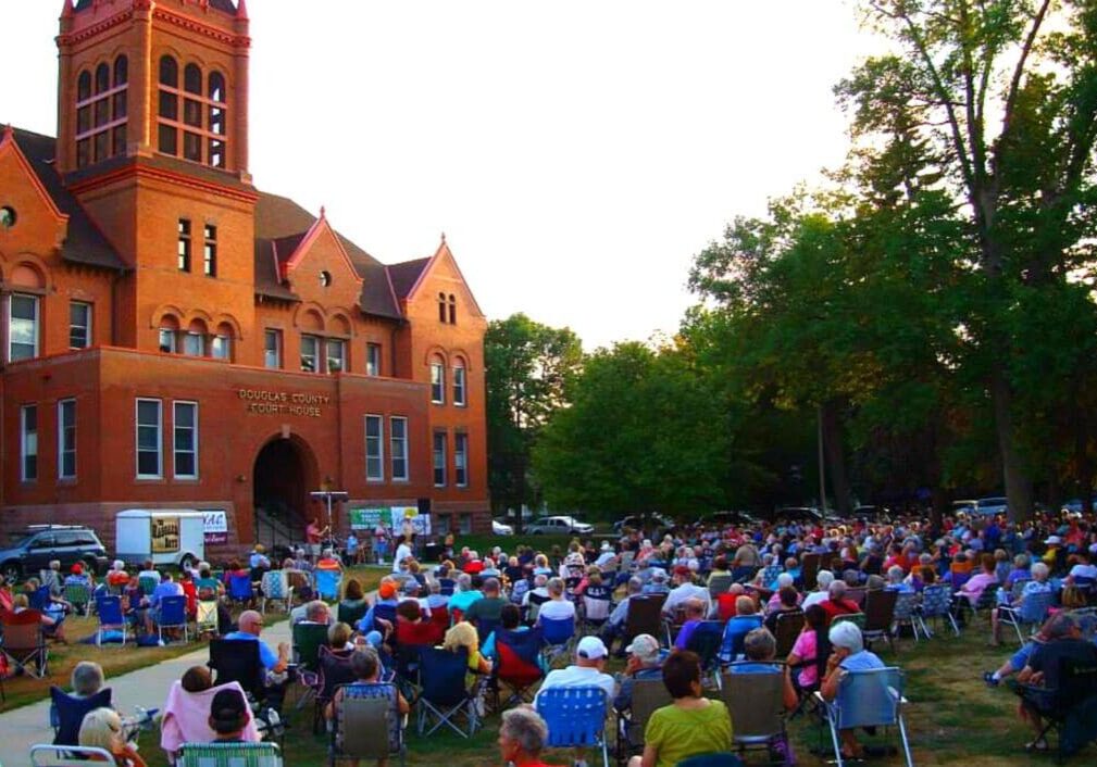 A large crowd sits on lawn chairs facing a stage set up in front of a historic red-brick courthouse, surrounded by trees, during Red Willow Arts’ lively outdoor community event at sunset.