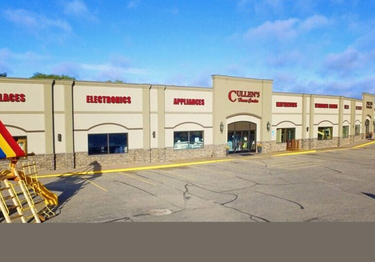A large retail store, Cullen's Home Center, displays signs for fireplaces, electronics, appliances, furniture, and mattresses. A wooden outdoor playset sits in the mostly empty parking lot beneath a clear blue sky.