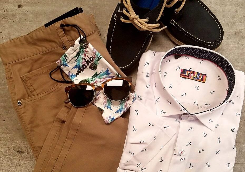 A flat lay of tan pants, a white shirt with small anchor prints, black boat shoes with tan laces, sunglasses, and a wallet with a tropical leaf pattern—perfect inspiration from The Dashery for your next downtown clothing shopping trip.