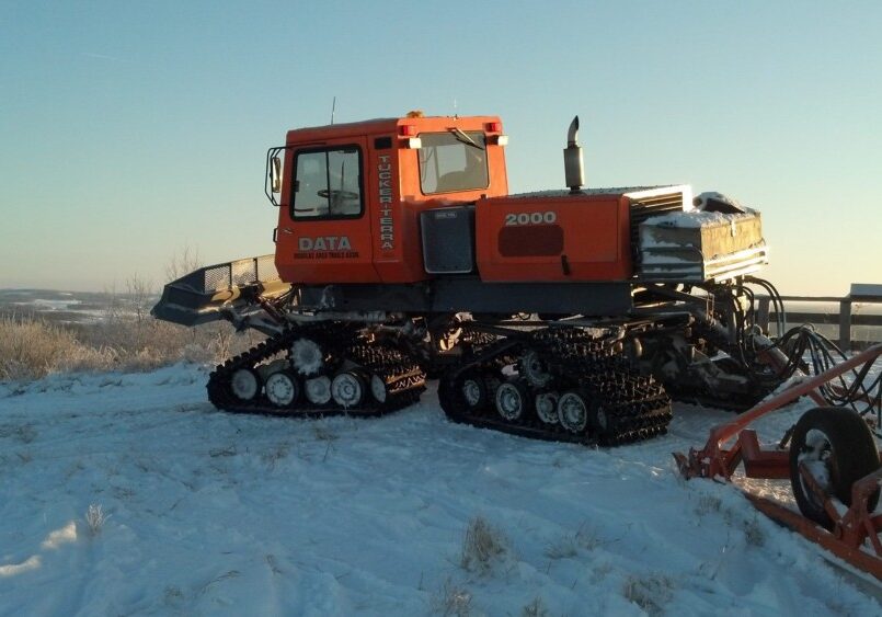 An orange snowcat vehicle with tracks, labeled "Tucker-Terra 2000," is parked on a snowy landscape under a clear sky, ready to maintain DATA Snowmobile Trails. Snow-covered ground and some equipment are visible around it.