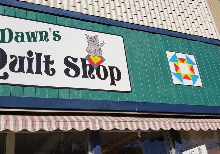 Dawn's Quilt Shop features a charming storefront sign with a gray cat holding a quilt and a vibrant geometric pattern. The shop’s green facade and striped awning invite guests into the cozy world of Dawn’s Quilt Shop.