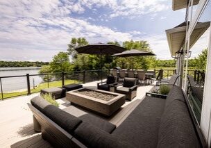 Spacious outdoor patio with modern black furniture, a fire pit, umbrellas, and a dining area—this Alexandria Dream Home overlooks a lake and is surrounded by lush greenery under a partly cloudy sky.
