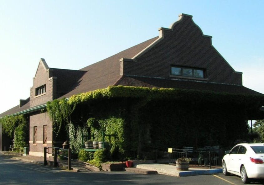 A brick building with a steep, gabled roof is partially covered in green ivy. Near Depot Smokehouse & Tavern, a white car is parked and outdoor tables and chairs line the shaded side of the building.