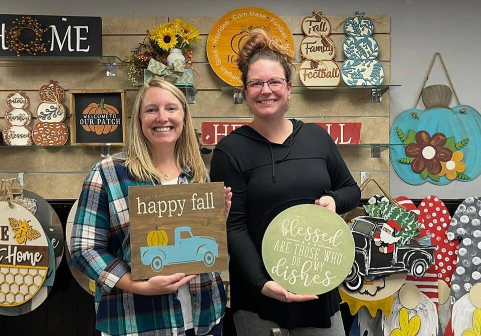 Two women smiling and holding autumn-themed wooden signs at Design Station; one sign says "happy fall" with a blue truck and pumpkin, the other says "blessed are those who do my dishes." Colorful fall decor hangs on the wall behind them.