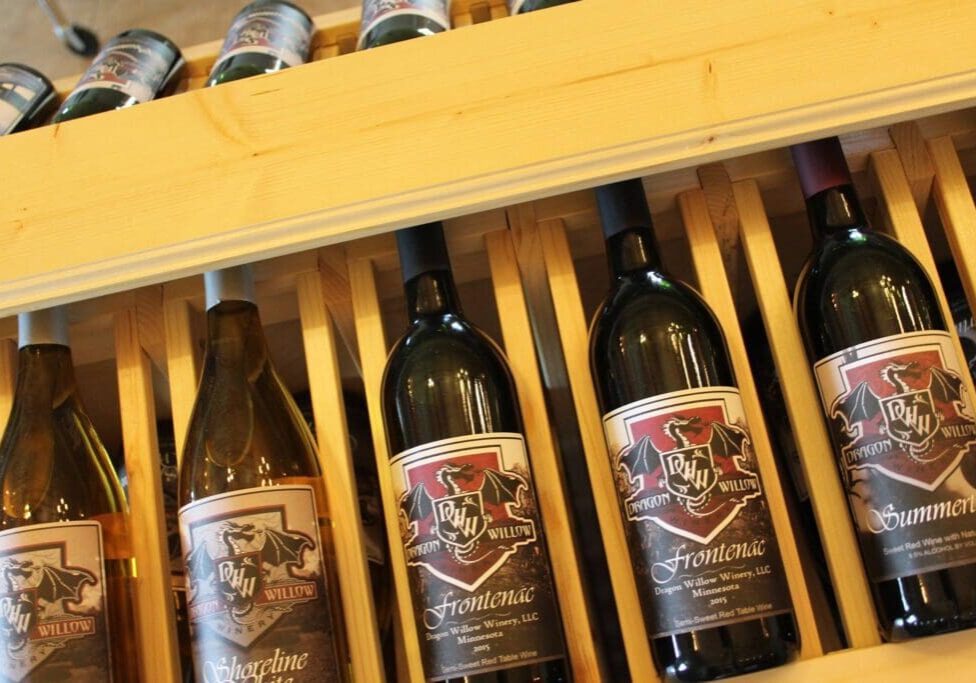 Several bottles of wine with decorative labels are displayed on a wooden rack. The bottles, including varieties like Frontenac, showcase the Dragon Willow Winery logo and are positioned side by side.