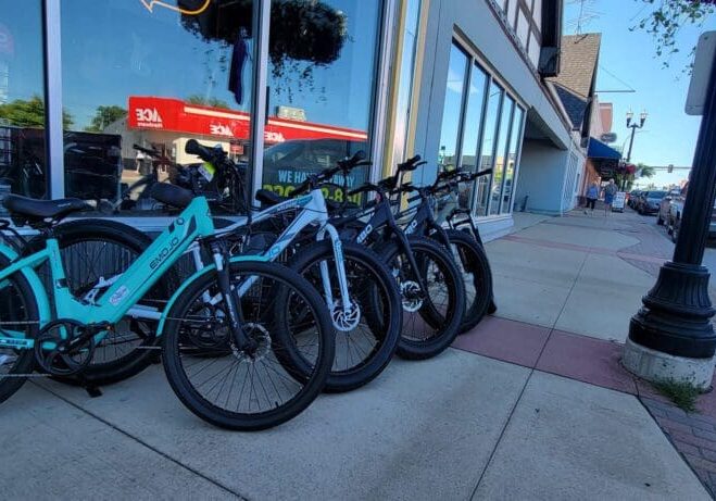 Several bicycles, including some from Central Ebikes, are lined up and parked on a sidewalk outside a storefront, with cars parked along the street and buildings visible in the background.