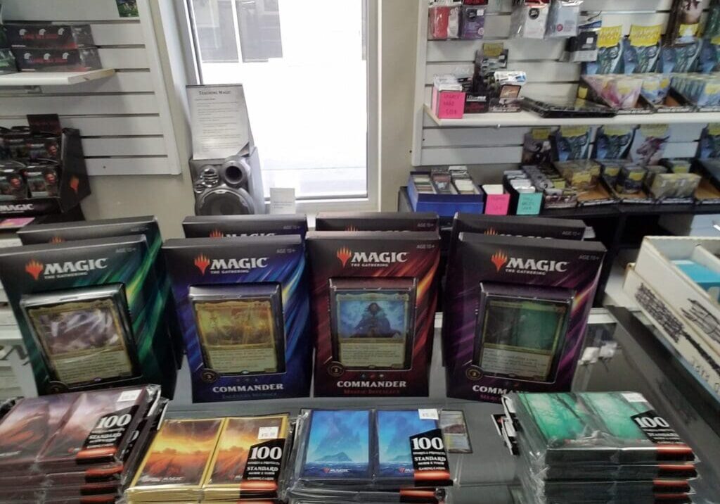 Four Magic: The Gathering Commander decks are displayed on a glass counter at Elemental Games, surrounded by card sleeves and packs, with shelves of gaming products visible in the background.