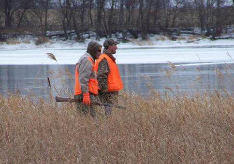 Two people wearing orange vests stand in tall brown grass near a partially frozen lake at Elk Lake Heritage Preserve, with one person holding a shotgun. Leafless trees line the background under a cloudy sky.