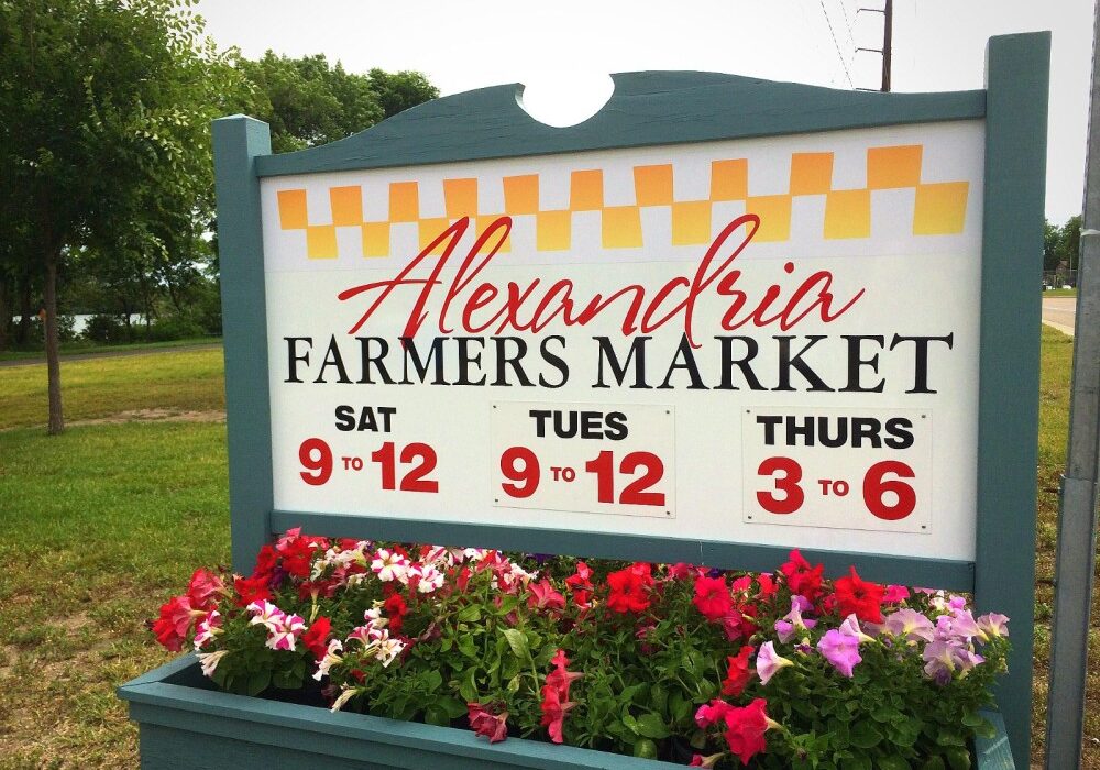 A sign for the Alexandria Farmers Market displays hours: Saturday and Tuesday from 9 to 12, and Thursday from 3 to 6. Colorful flowers bloom at its base, with trees and grass surrounding the vibrant Alexandria Farmers Market scene.