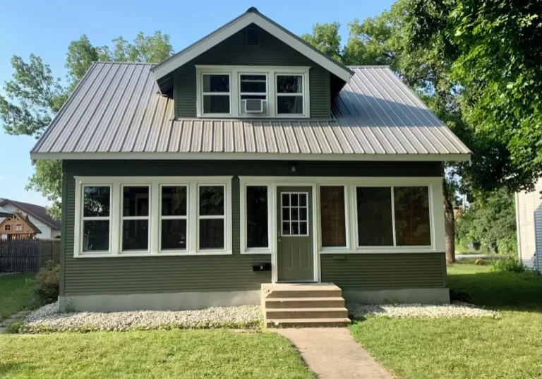 The Boho Bungalow is a green two-story house with a gray metal roof, large front windows, and a small front porch with steps, set on a grassy lawn with trees and a sidewalk leading to the door.