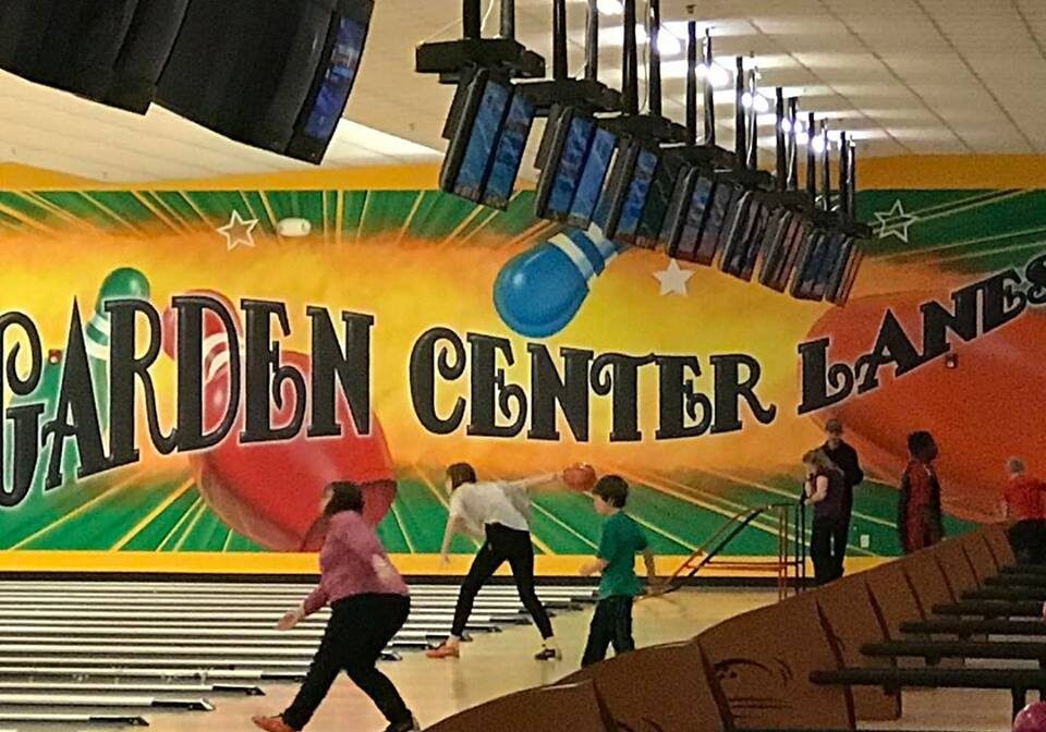 Several people, including children, are bowling at Garden Center Lanes, an indoor alley with colorful wall art and multiple monitors hanging above the lanes.