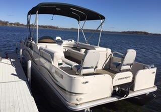 A white pontoon boat with a canopy, by Garfield Marine, is docked at a wooden pier on a calm lake under a clear blue sky. The boat has seating areas and the word "Sanpan" visible on the back.