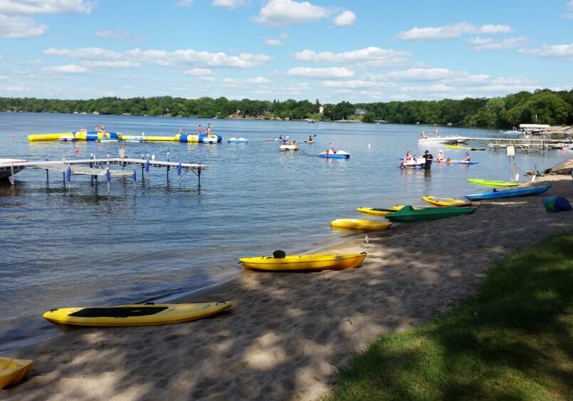 A sandy lakeshore at Geneva Beach Resort features colorful kayaks, docks stretching into the water, and people swimming and playing on floating inflatables under a sunny sky. Trees line the distant shore of the lake.