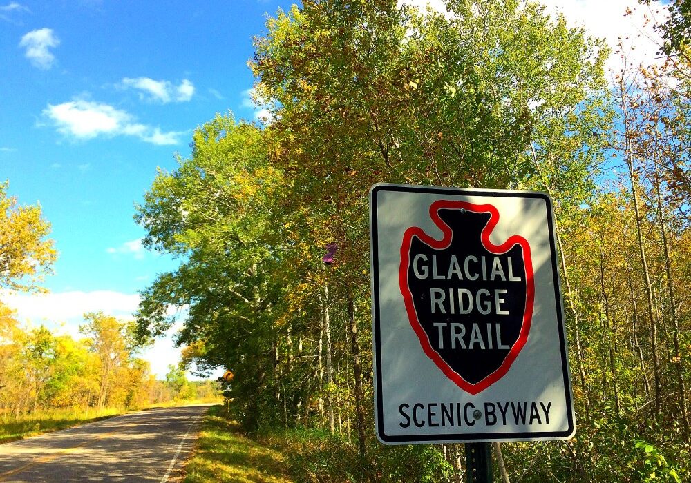 A road sign marks the Glacial Ridge Trail Scenic Byway beside a rural road, framed by green and yellow trees beneath a bright blue sky dotted with clouds.