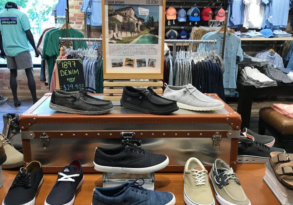 A display of men’s casual shoes on a vintage trunk at Glik's clothing store, with shirts and hats on racks in the background. A sign featuring a photo and story is showcased above the shoes.