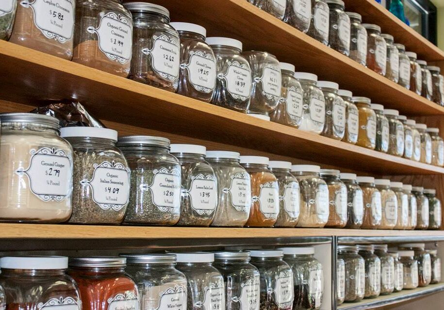 Shelves at The Grain Bin are filled with glass jars containing various herbs, spices, and seasonings, each labeled with the name and price of the contents. The jars are organized neatly in rows.