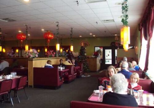 A casual Chinese restaurant, Great Hunan features red booths, hanging lanterns, and several diners seated at tables. The atmosphere is cozy with natural light streaming through large windows on the right side.