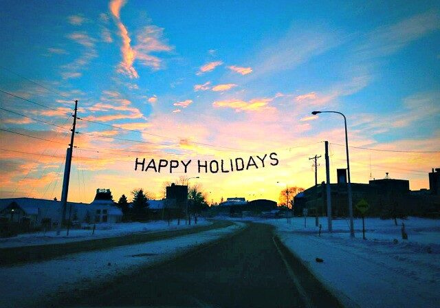 A snowy street in Alexandria at sunset, with buildings on either side and a "HAPPY HOLIDAYS" banner stretched across the road. The colorful sky and festive scene invite all to celebrate the season amid blue, orange, and pink clouds.