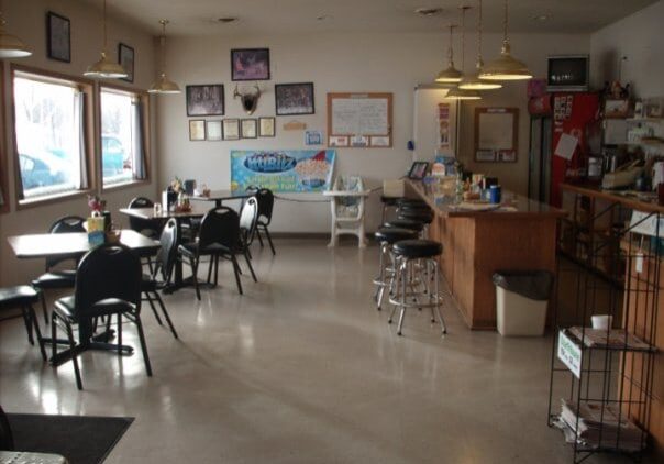 Casual diner interior at Herby's with tables and chairs, a counter with barstools, large windows, wall decorations, and a refrigerator in the corner. The space appears clean and well-lit with natural and artificial light.