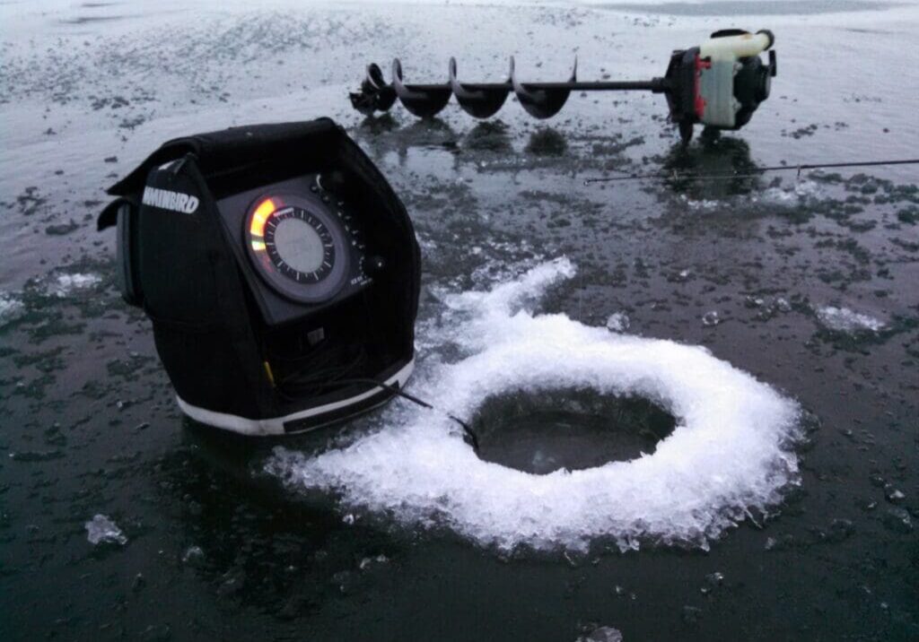 An ice fishing sonar sits next to an icy hole drilled in a frozen lake, helping anglers monitor ice depths in Alexandria area waters, with an ice auger lying nearby on the snowy surface.