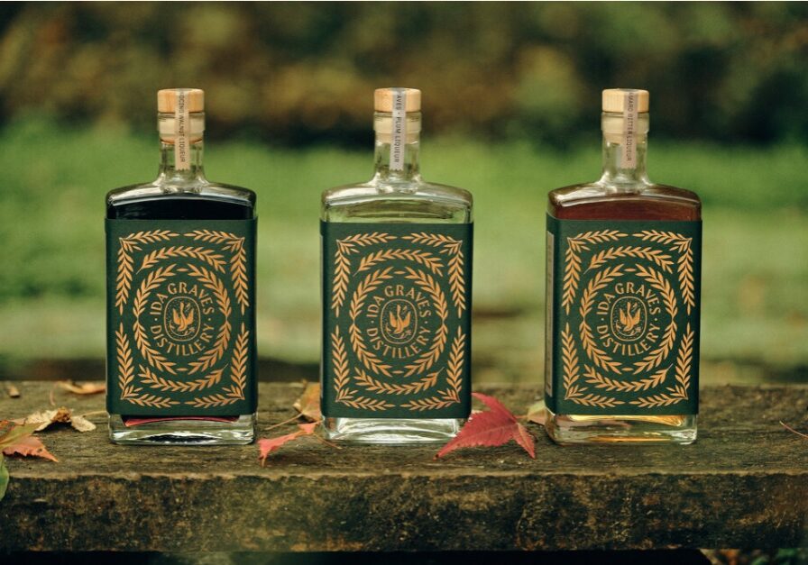 Three bottles of spirits with green and gold labels reading "MS GRANS DISTILLERS" from Ida Graves Distillery are displayed on a rustic wooden surface outdoors, autumn leaves scattered nearby and a blurred green background.