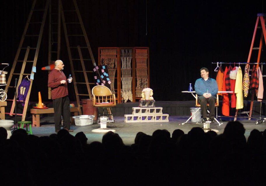 Two actors perform on stage surrounded by props, costumes, and household items, with a seated audience watching. As they engage in dialogue, their scene humorously bids 2014 farewell and captures the festivity of New Year’s Celebrations.