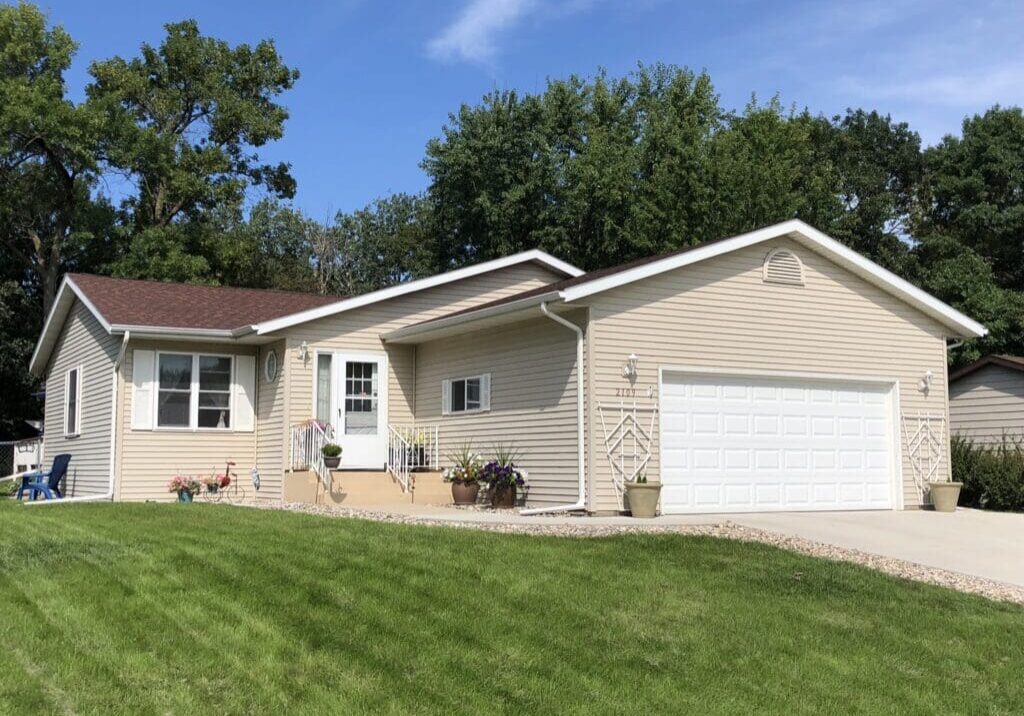 Into the Blue Vacation Home is a single-story beige house with a red-brown roof, white garage door, front porch with railings, potted plants, and a well-kept green lawn, set against trees and a blue sky.
